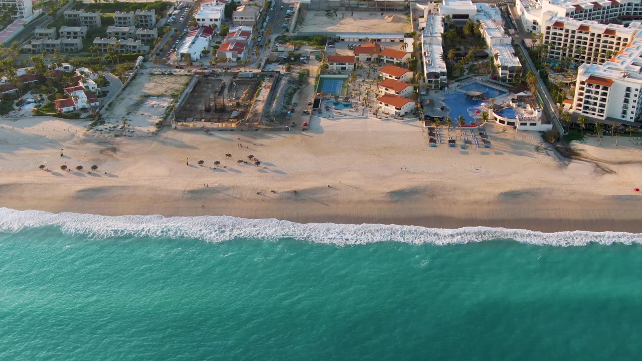 Aerial frame over San Jose Del Cabo showing turquoise water, a thin white surf line, pale sand, resort pools, and hotel buildings with no visible people.