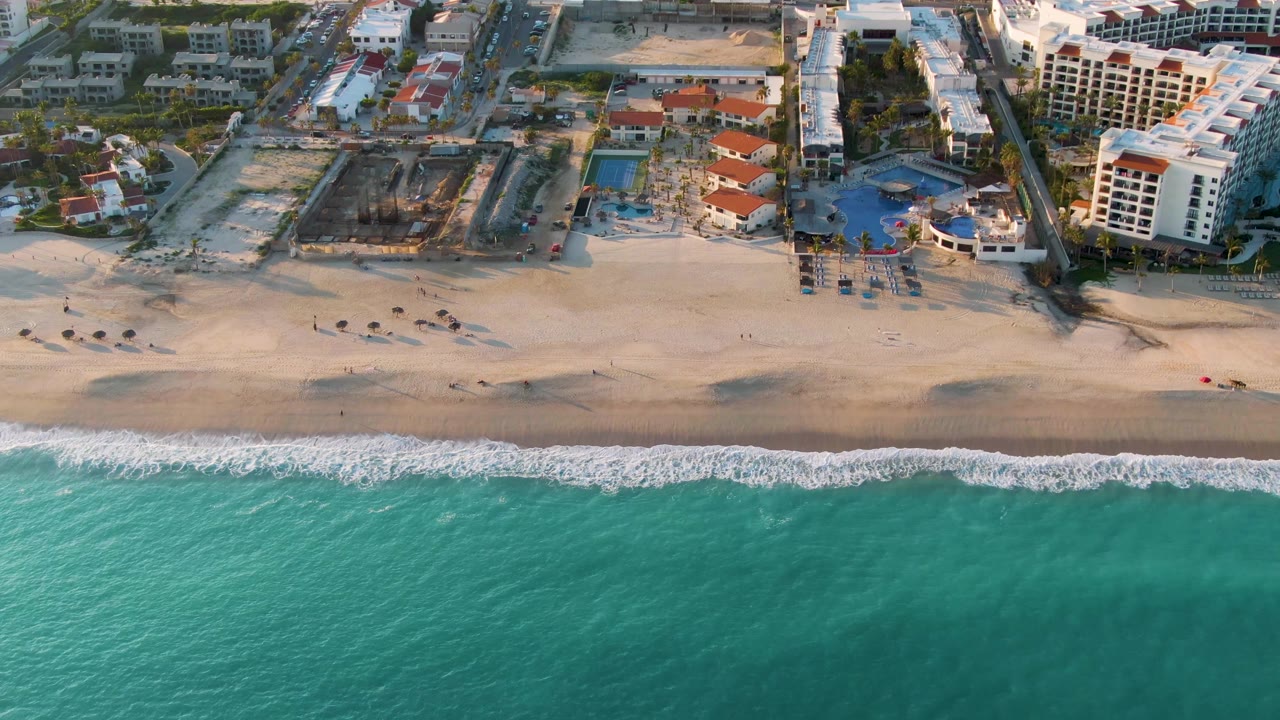 San Jose Del Cabo beachfront from above, with the ocean lower in frame, waves nearing the sand, a blue pool area, and resort blocks along the shore with no visible people.