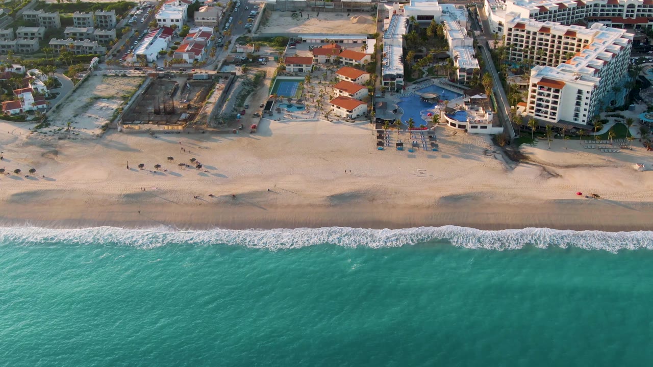 A later aerial frame of San Jose Del Cabo beach shows a broad turquoise water band, foamy shore break, resort pools, and multi-story buildings with no visible people.