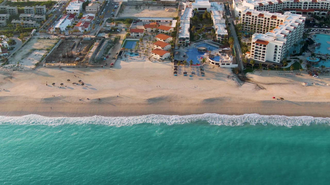 Aerial coastline view with rolling waves, empty-looking sand, and resort pools.