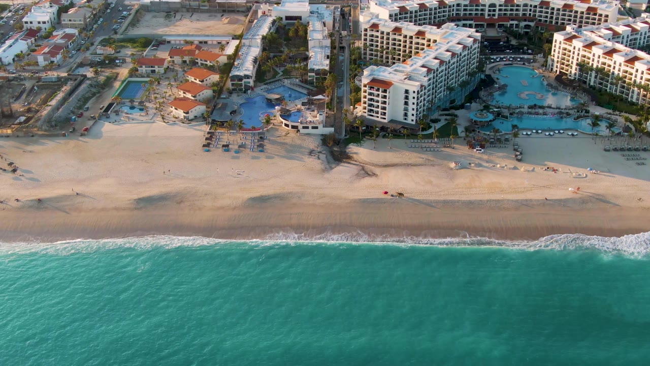 Aerial view in San Jose Del Cabo with foamy surf slightly farther along the shoreline, hotel wings and swimming pools behind the beach, and no visible people.