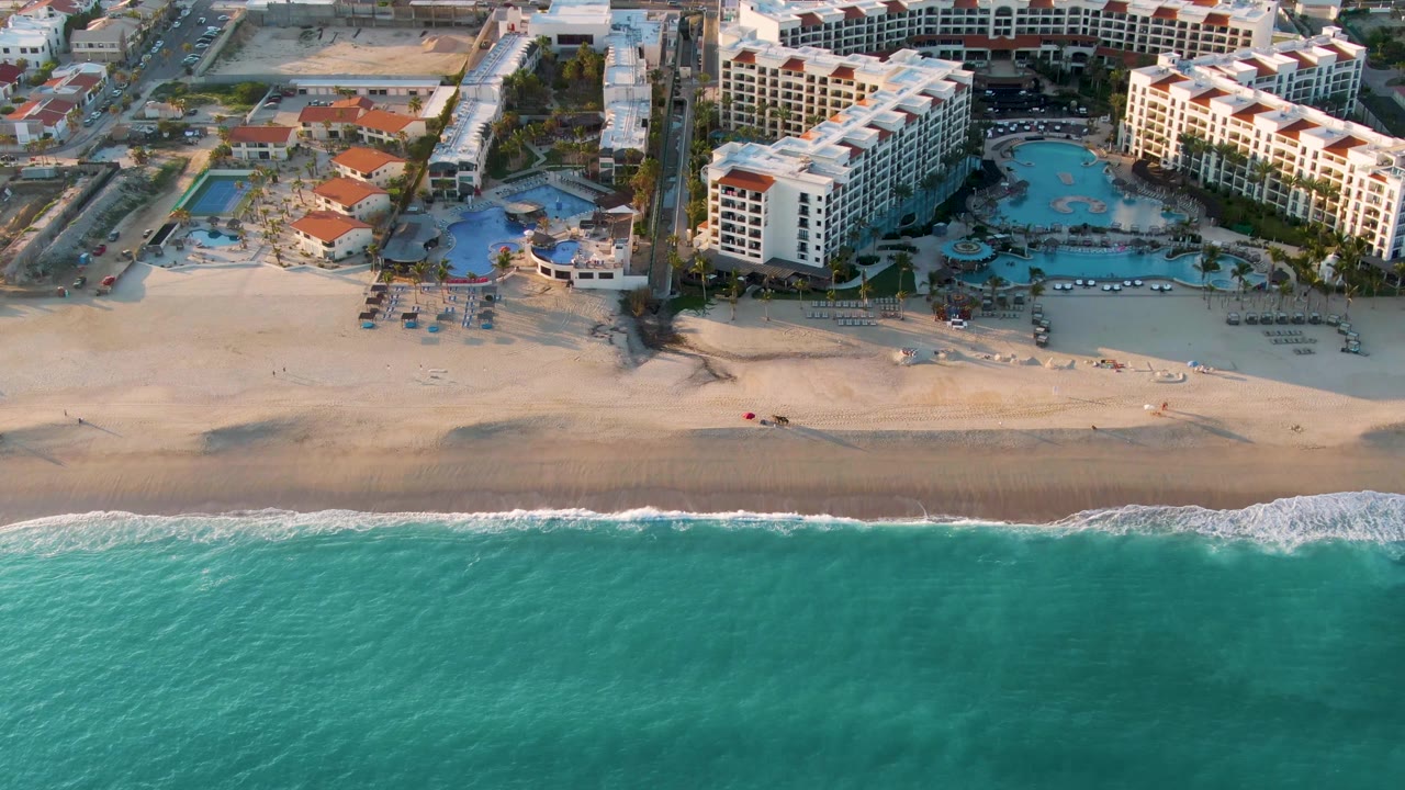 Aerial frame from San Jose Del Cabo showing the turquoise water band below a pale beach, with white hotel buildings, pool areas, and no visible people.