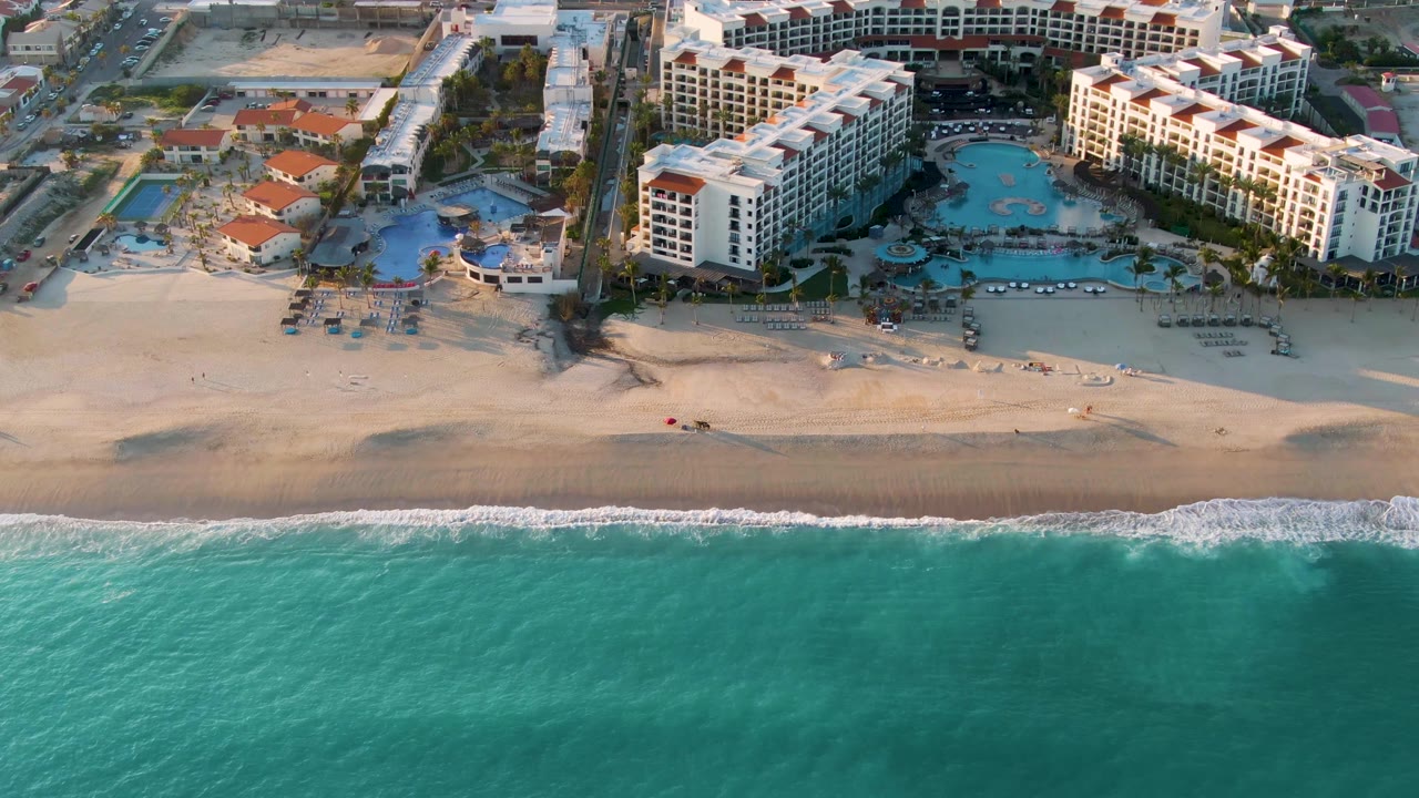 Mid-clip aerial frame in San Jose Del Cabo showing the resort pool courtyards, white beachfront buildings, broad sand strip, shifting foam, and no visible people.