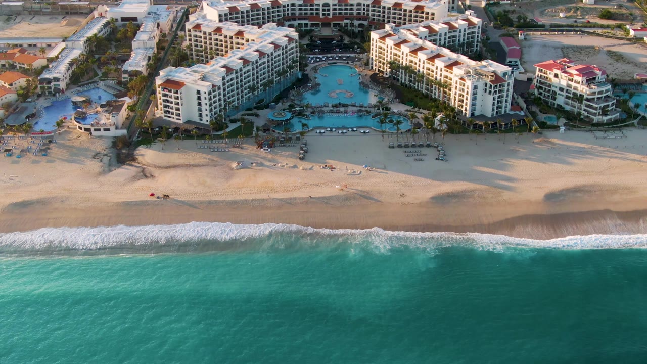 At 0:01, the San Jose del Cabo aerial view holds on the resort pools and beachfront, with waves crossing the lower frame and no visible people.