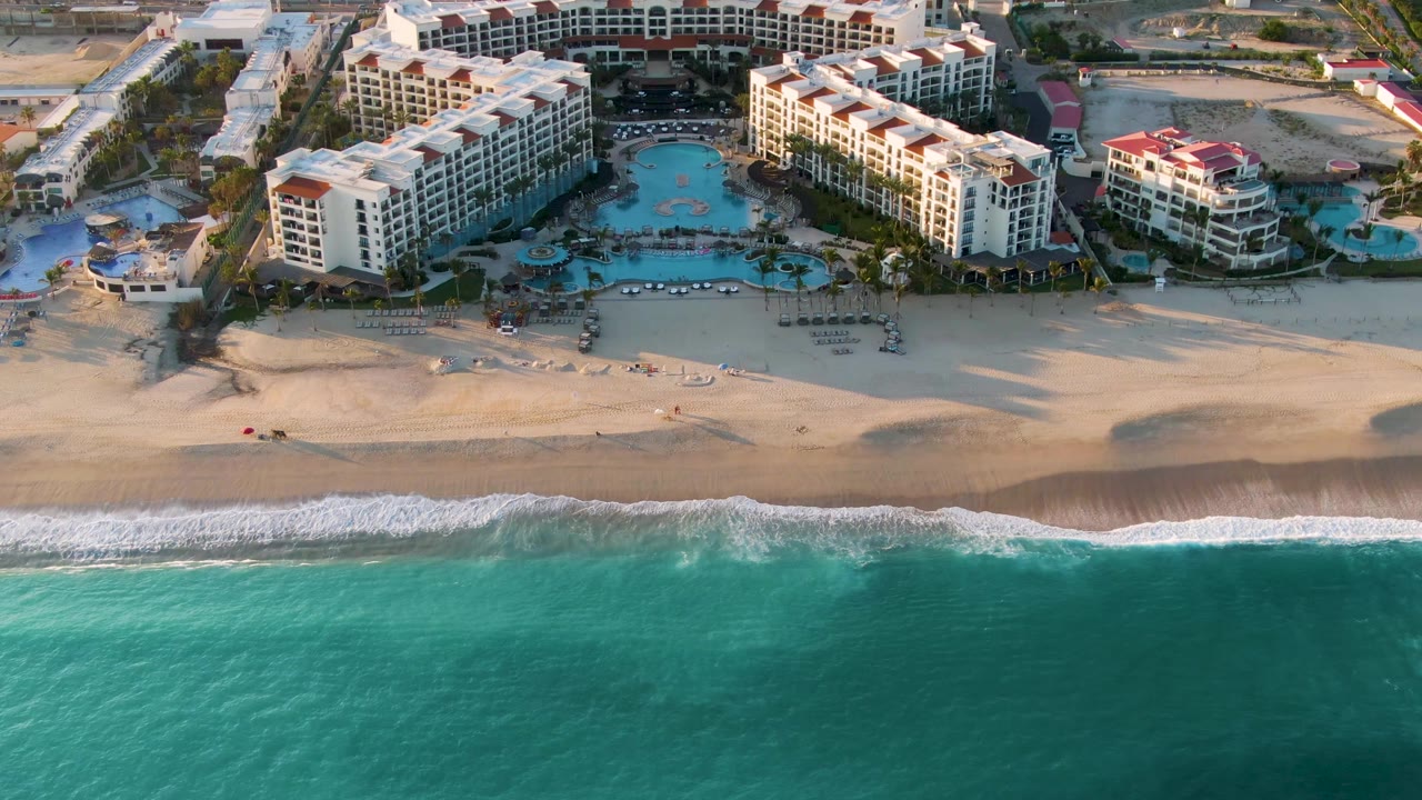 At 0:02, the drone angle shows more of the resort's right side in San Jose del Cabo, with white buildings, blue pools, beach sand, and no visible people.