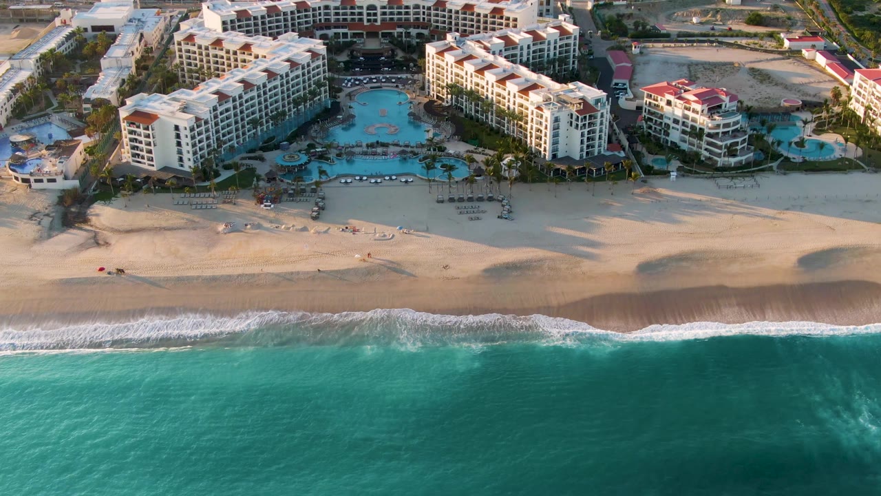 At 0:03, the aerial view centers the beach and pool area, with the turquoise ocean at the bottom edge and San Jose del Cabo resort buildings beyond, with no visible people.