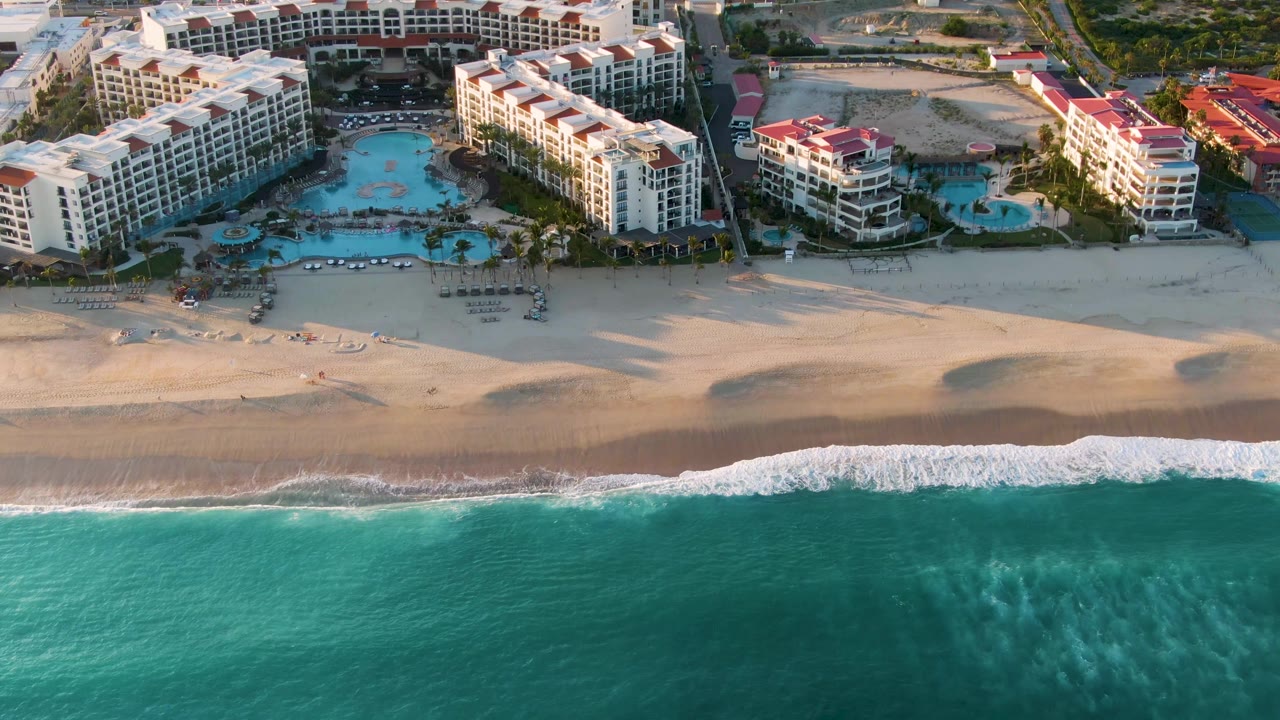 Aerial frame from San Jose Del Cabo showing turquoise water below a sandy beach and resort buildings with pools behind the shore, with no visible people.