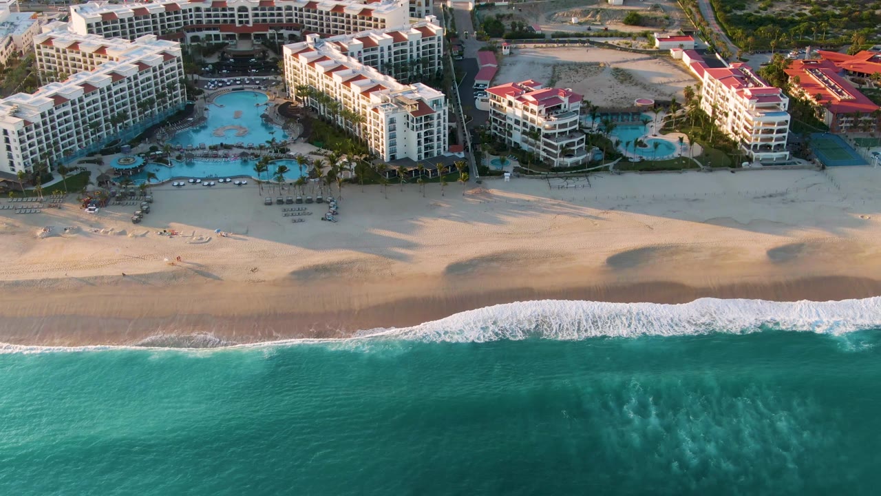 Aerial view in San Jose Del Cabo with the white surf line slightly higher on the sand, turquoise ocean in front, and resort pools and buildings in the background, with no visible people.