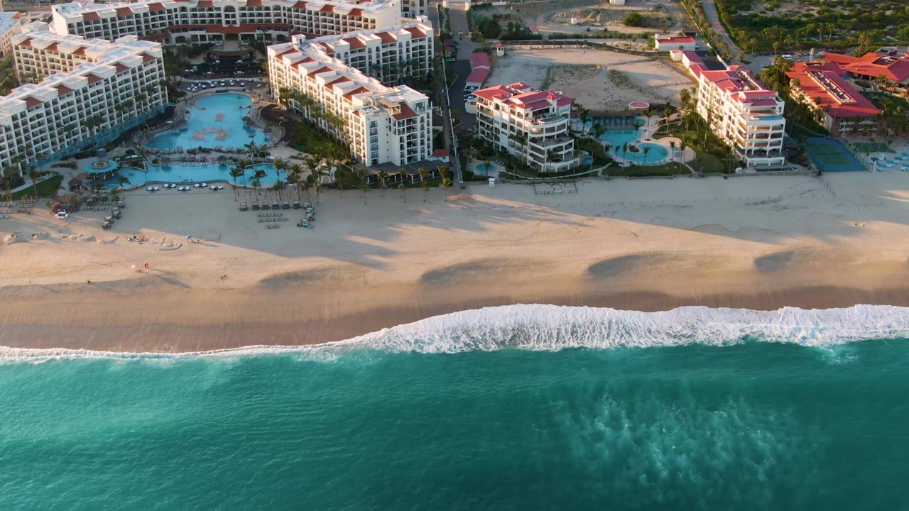 San Jose Del Cabo aerial beach frame with white foam stretching along the shoreline, darker wet sand beneath it, and resort buildings and pools beyond the beach, with no visible people.