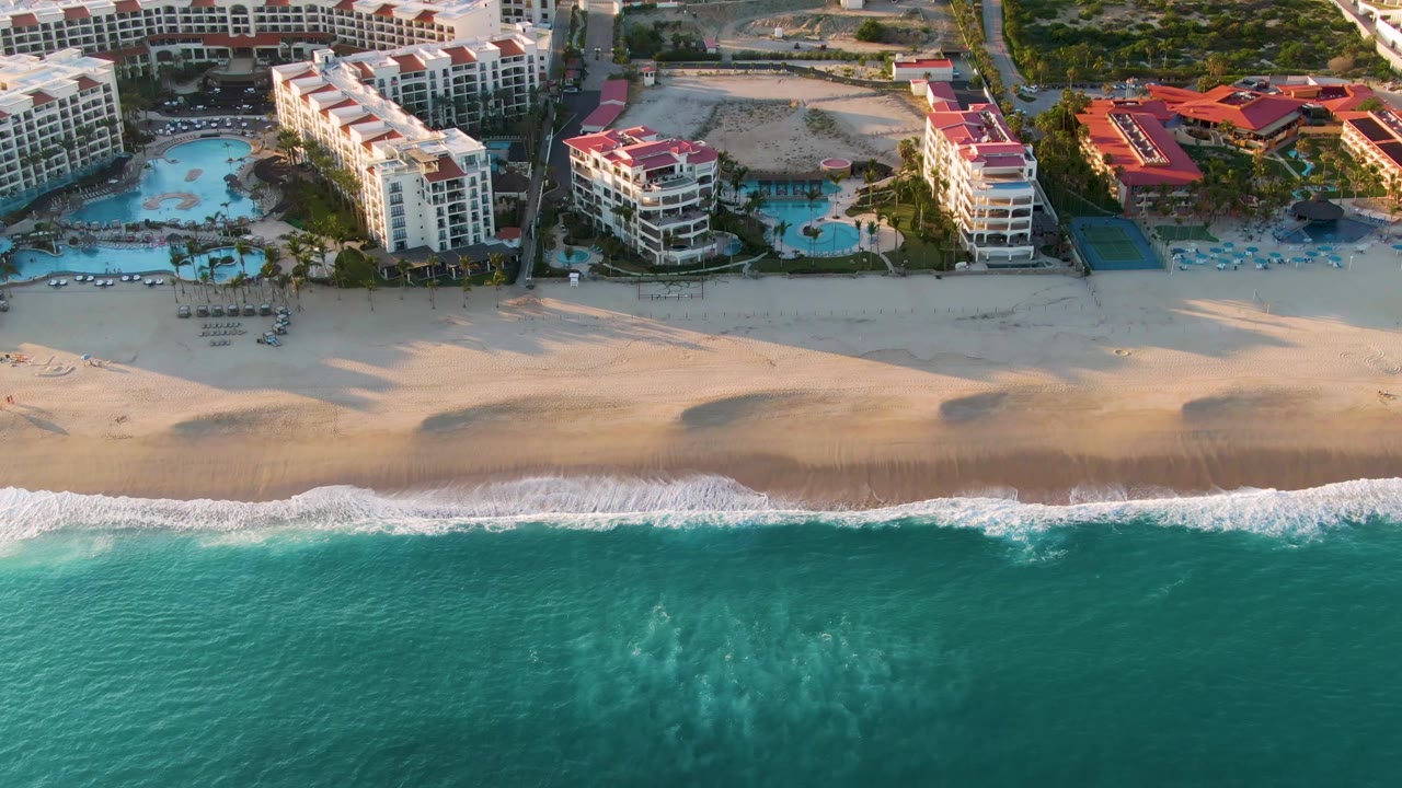 High aerial view over San Jose Del Cabo showing turquoise water, a wide sandy beach, and white resort buildings with red roofs, with no visible people.