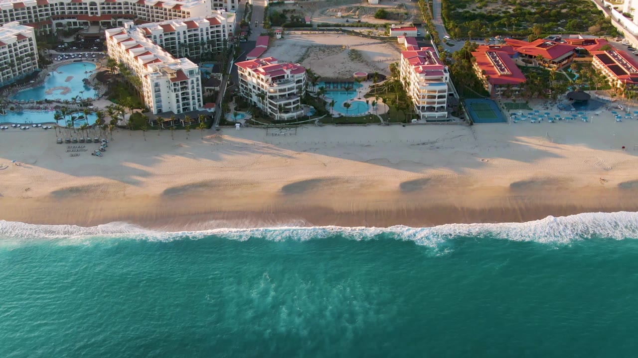 Aerial frame in San Jose Del Cabo with a broad band of teal ocean, a thin line of breaking surf, open sand, and resort blocks set back from the beach, with no visible people.