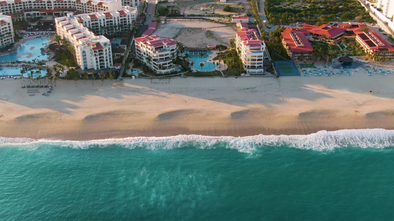 San Jose Del Cabo aerial view with foamy waves spreading along the beach, pale sand in warm light, and resort buildings and pools across the upper frame, with no visible people.