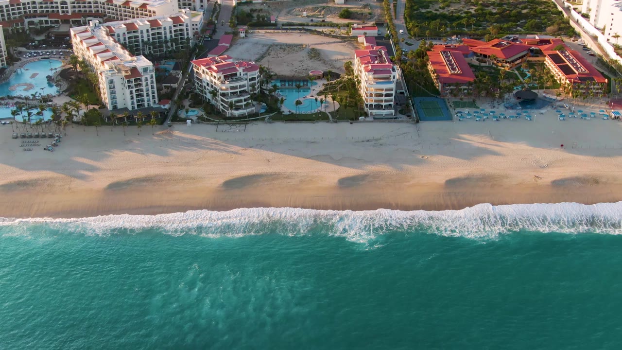 Aerial view of resort beachfront with waves, sand, pools, and hotel buildings.