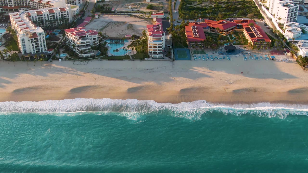 Aerial view in San Jose Del Cabo, Mexico, with turquoise ocean, a white foam line, broad sand, red-roof resort buildings, pools, boats, and no visible people.