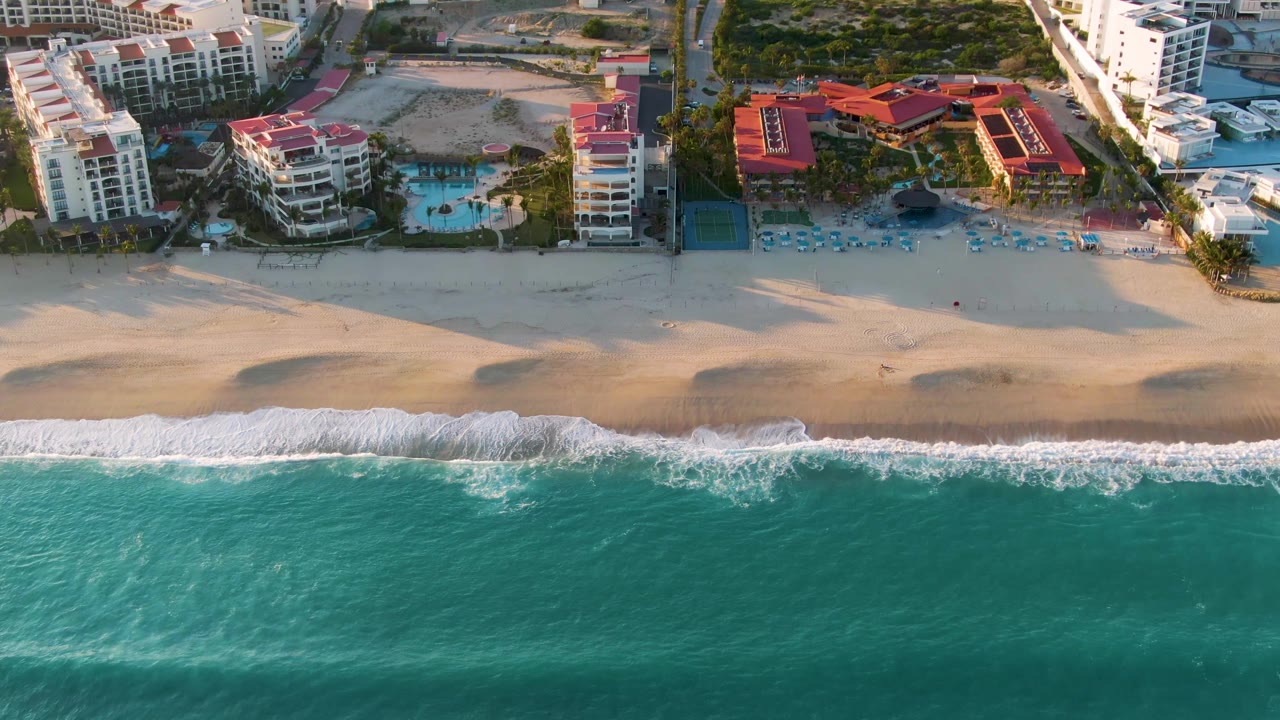 San Jose Del Cabo aerial frame showing the surf line slightly farther up the sand, with red-roof resort structures, pool areas, boats, and no visible people.