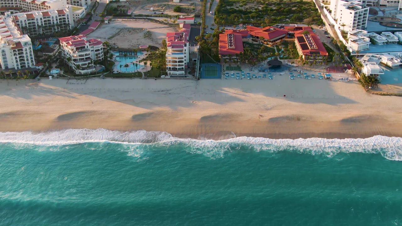 Aerial San Jose Del Cabo coastline frame with a wide turquoise water field, broken white surf, pale sand, beachfront resort buildings, pools, boats, and no visible people.