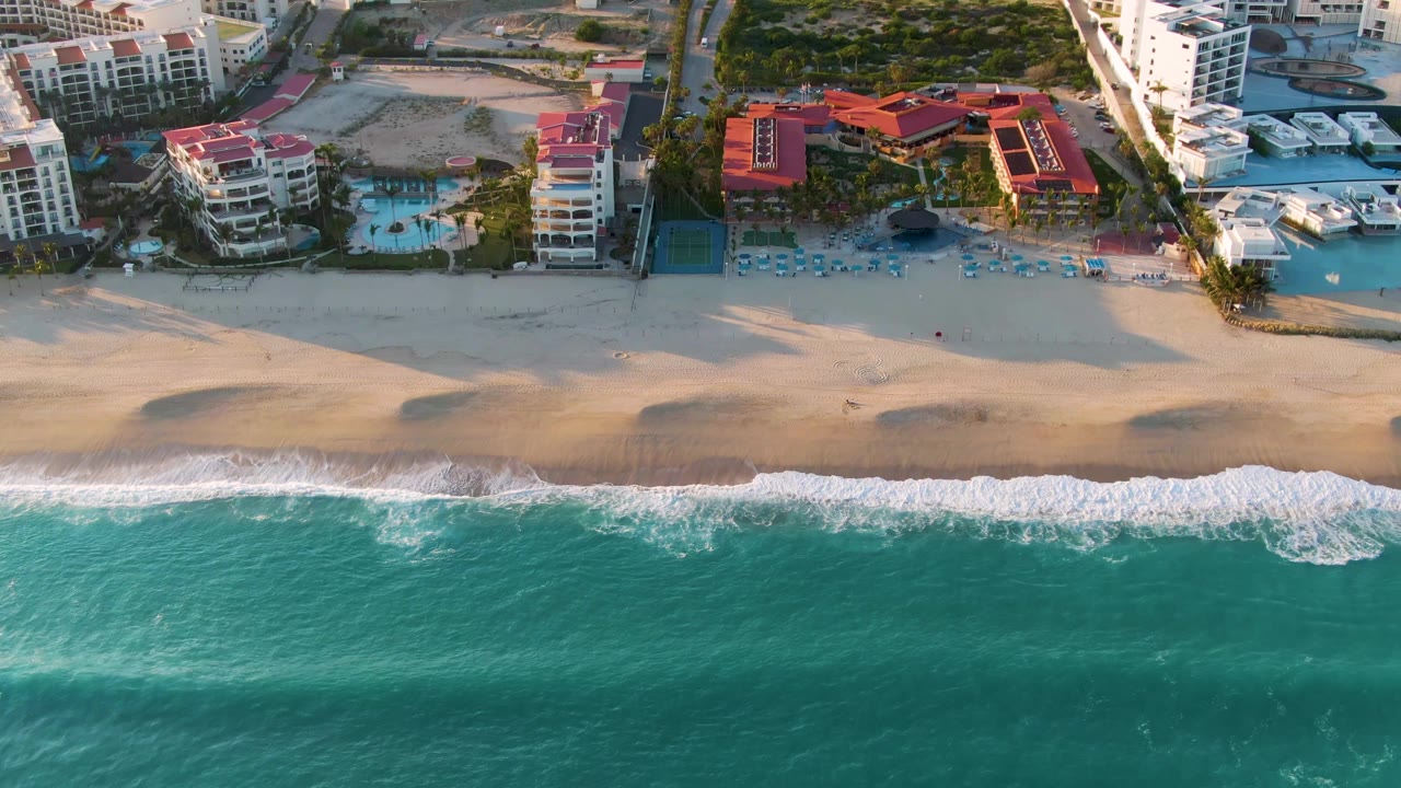 San Jose Del Cabo aerial view where the foam spreads across the lower edge of the beach, with red-roof buildings, pool courtyards, boats, and no visible people.