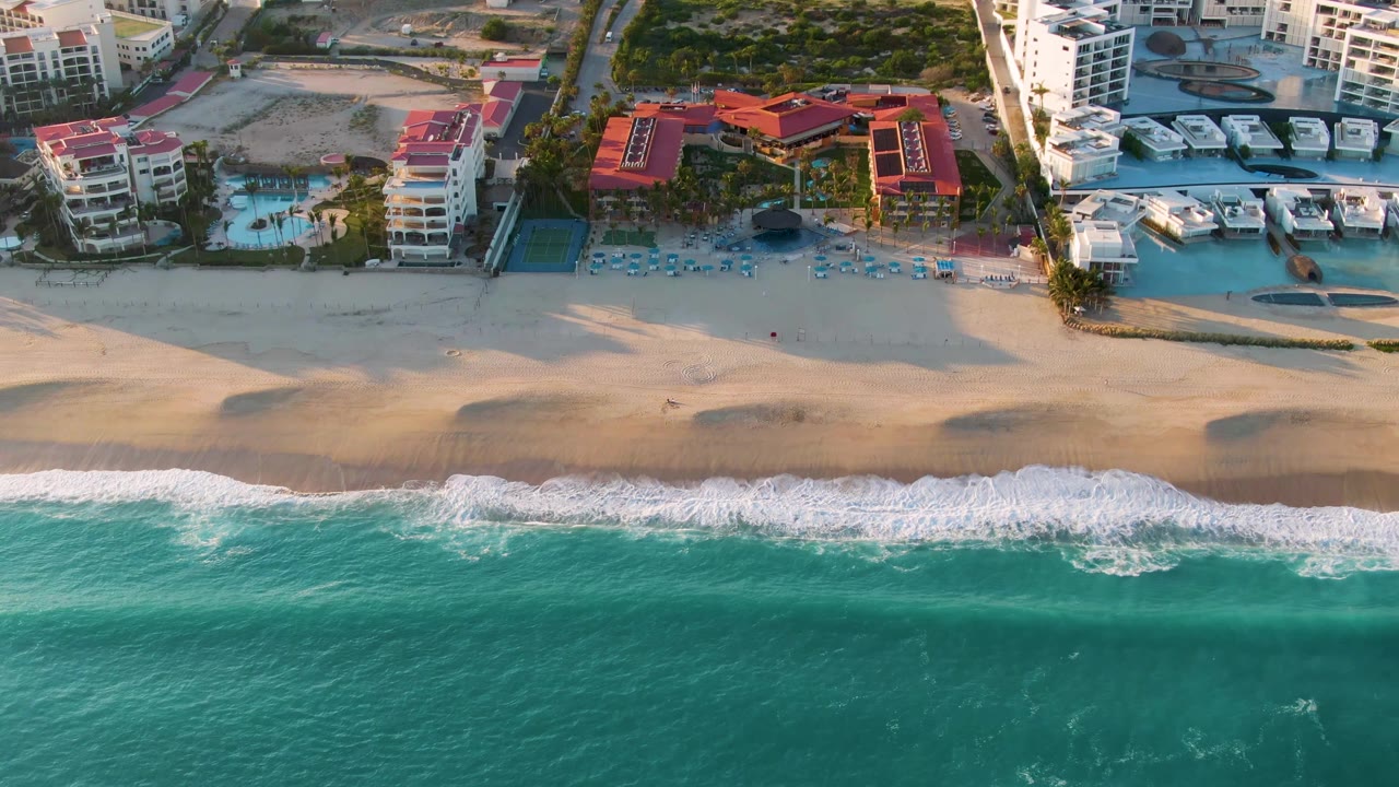 Aerial view in San Jose del Cabo showing turquoise ocean, white surf, wide sand, beachfront buildings, pools, and boats at the start of the clip, with no visible people.