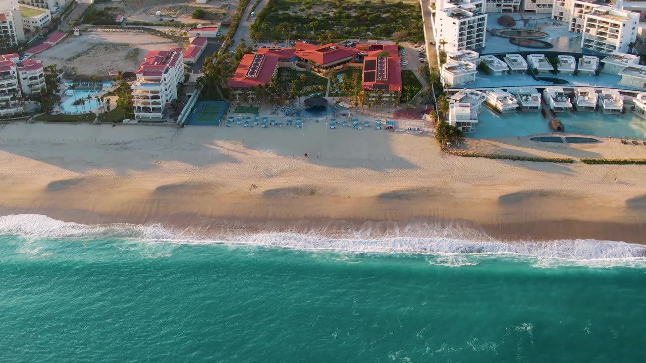 Drone-style aerial view in San Jose del Cabo with a broad sandy beach between green-blue water and coastal buildings, while the surf pattern shifts along the shore, with no visible people.