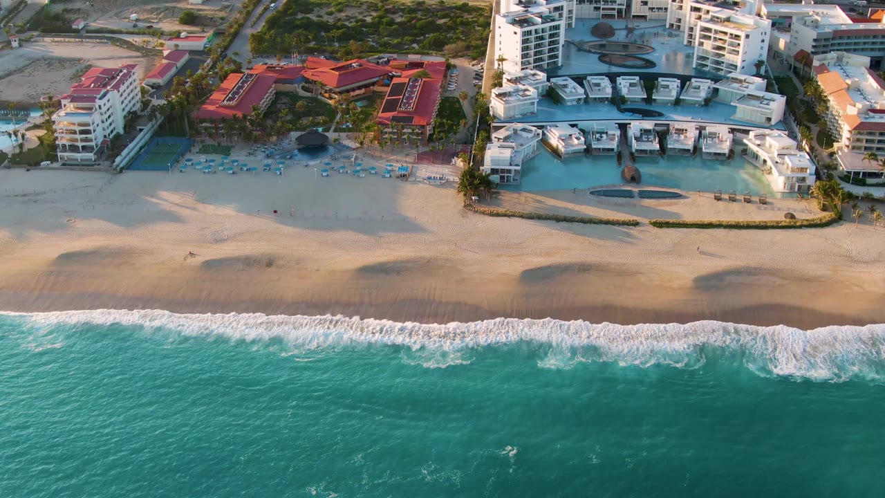 Aerial frame over San Jose Del Cabo shoreline with turquoise water, a band of white surf, tan beach, blue pools, red roofs, and white resort buildings, with no visible people.