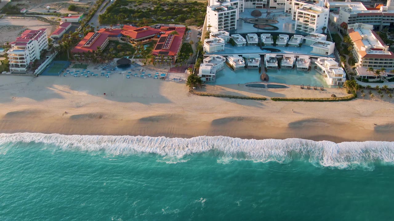 Drone view of beachfront resort pools behind a sandy beach and breaking waves.