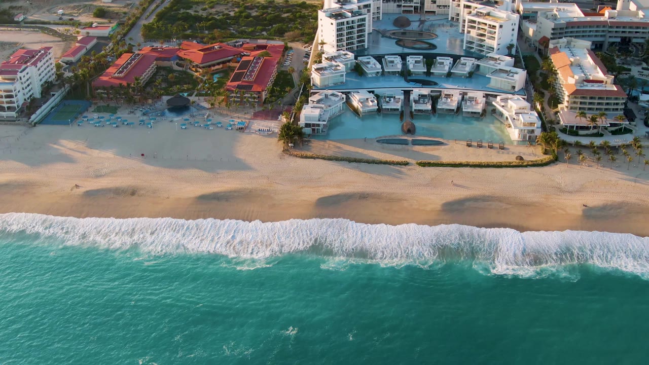 Aerial view along the San Jose Del Cabo beach with turquoise ocean filling the lower half, white waves reaching the sand, and resort buildings and red roofs above the shoreline, with no visible people.