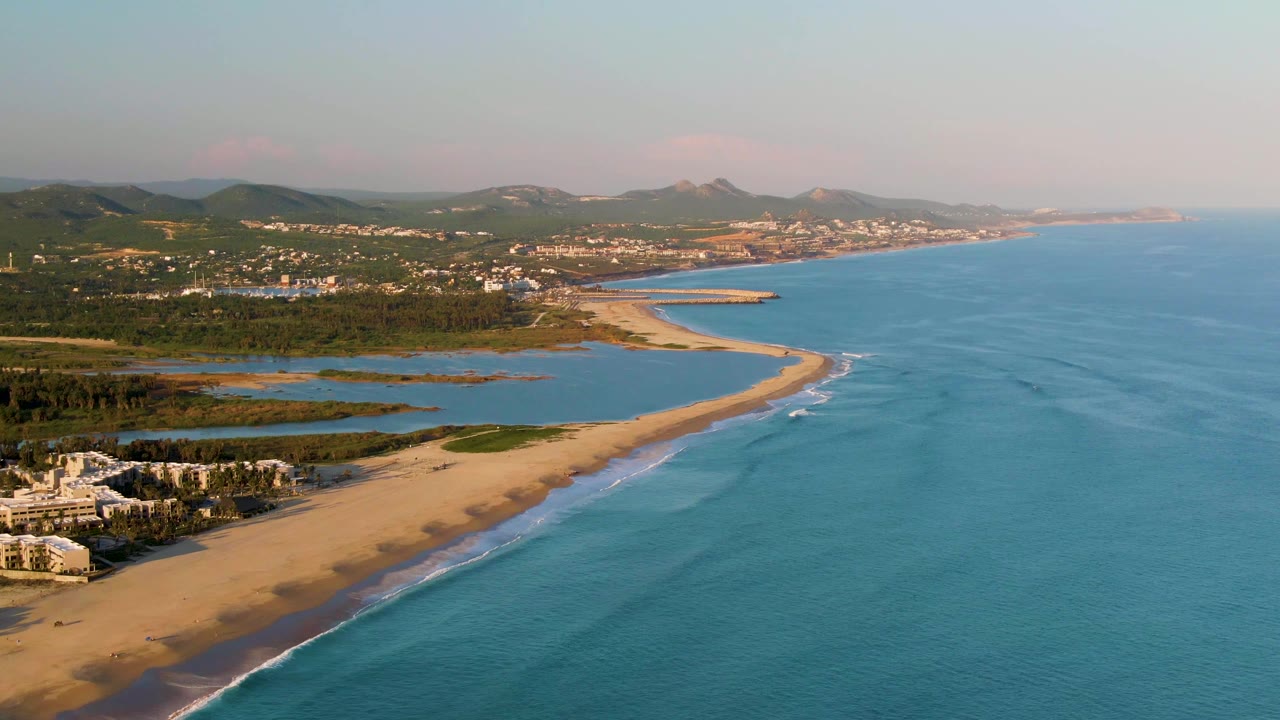 Aerial coastline with sandy beach, turquoise ocean, inland water, greenery, buildings, and distant hills.
