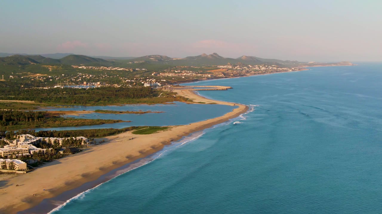 Aerial frame in San Jose del Cabo showing small waves curling onto the long sandbar-like beach, blue ocean filling the right side, green inland areas and low hills beyond, with no visible people.