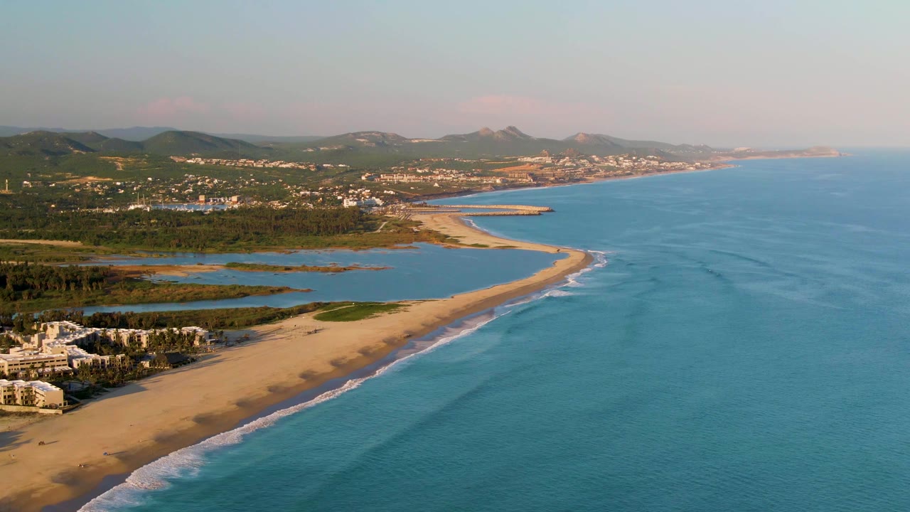 Mid-clip aerial frame over San Jose del Cabo coastline with the sandy beach running across the lower half, calm blue ocean to the right, green land and water channels inland, distant town buildings, and no visible people.