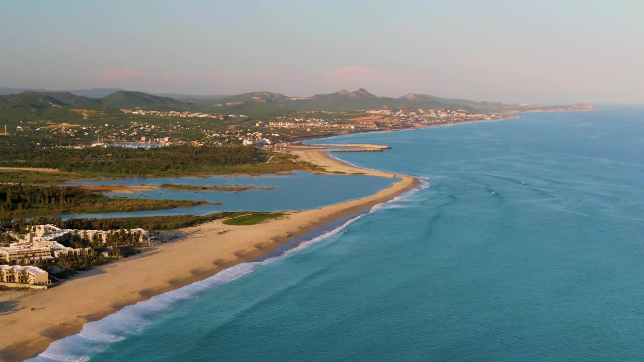Wide aerial frame of San Jose del Cabo coastline with a sandy beach curving beside blue ocean, inland water, distant hills, scattered buildings, and no visible people.