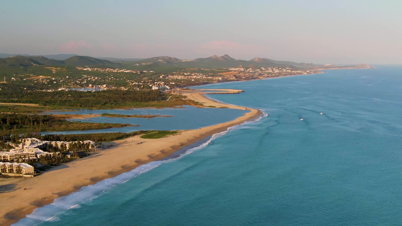 Aerial frame over San Jose del Cabo beach with turquoise-blue water filling the right side, a long strip of sand, small boats offshore, distant hillside buildings, and no visible people.