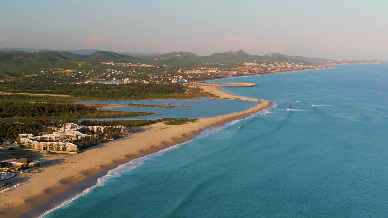 Aerial view in San Jose Del Cabo, Baja California Sur, showing a sandy beach beside turquoise ocean, a coastal lagoon, white shorefront buildings, green hills, and no visible people.