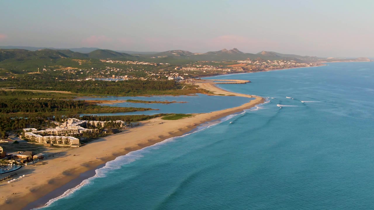 Wide aerial frame over San Jose Del Cabo coastline with the diagonal sandy shore, lagoon water behind it, small white wave lines offshore, clustered buildings, and no visible people.