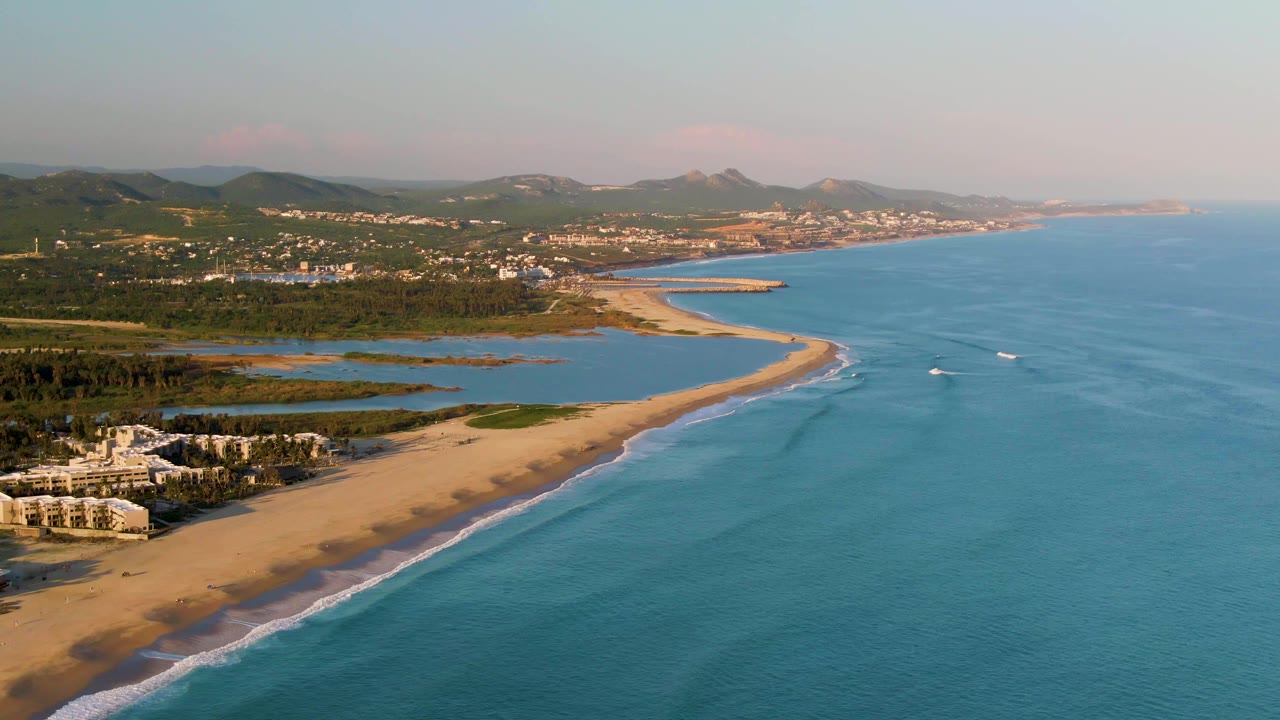 Opening aerial frame over San Jose del Cabo beach with blue ocean on the right, sandy shore below, distant hills, shoreline buildings, and no visible people.