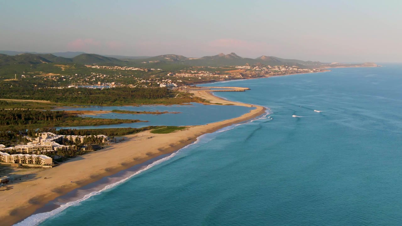 Wide aerial coastline with sandy beach, calm ocean, two boats, and hills.