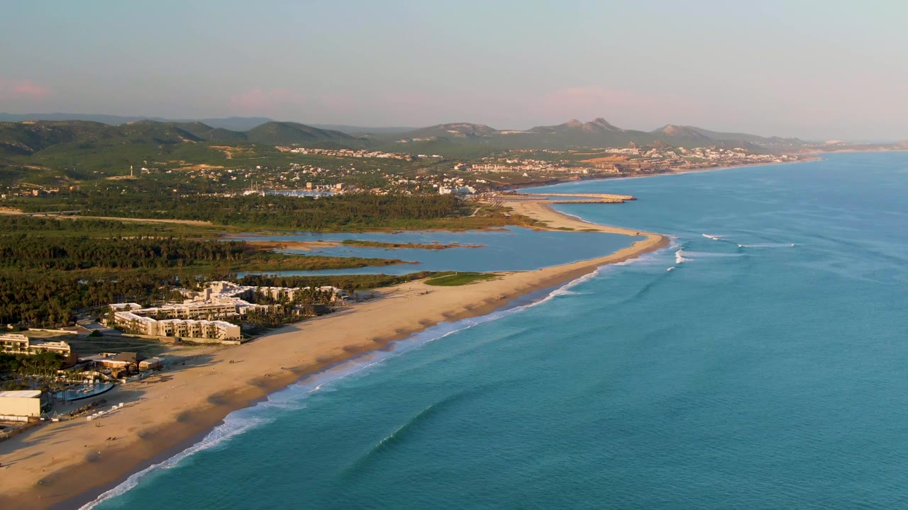 Aerial frame over San Jose del Cabo showing a sandy beach curving beside blue ocean, resort buildings in the lower left, inland water beyond the sand, distant hills, and no visible people.