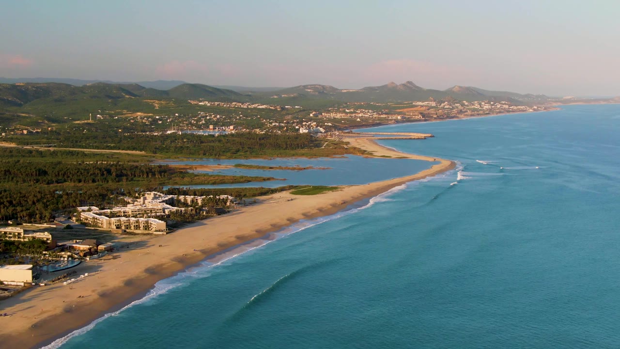 Aerial view in San Jose del Cabo with wave lines moving closer to the sandy shoreline, resort buildings at lower left, blue-green water to the right, inland wetlands, hills, and no visible people.