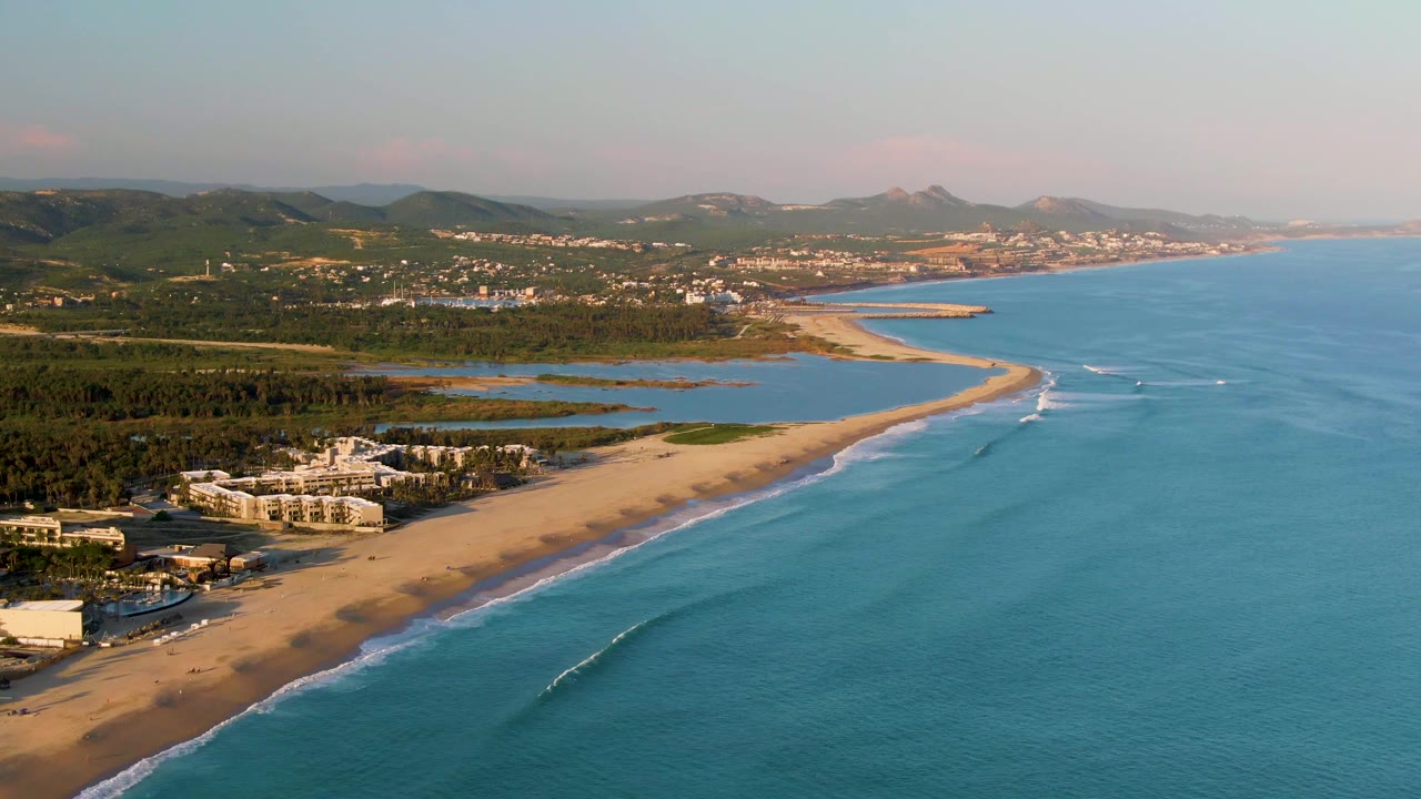 San Jose del Cabo aerial still with the sandy beach running diagonally through frame, turquoise water and a small breaking wave on the right, lagoon areas inland, distant hills, and no visible people.