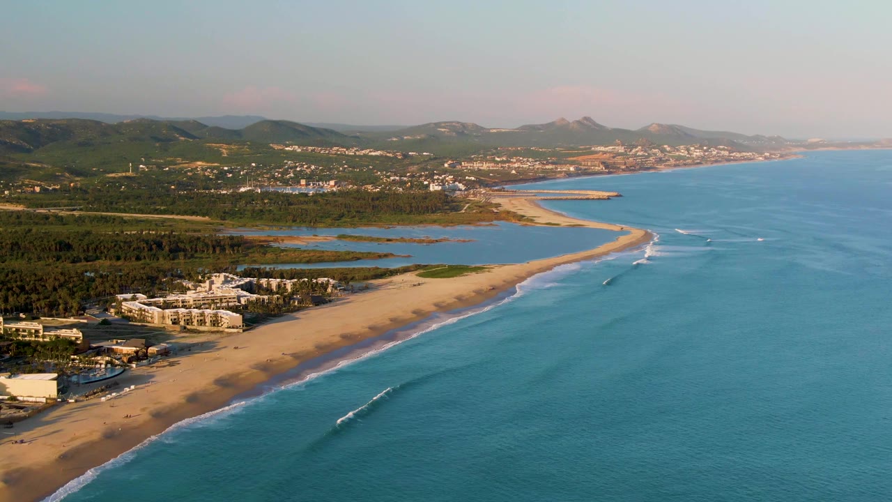 Drone-height view of resort buildings beside a sandy beach with blue water and distant hills.