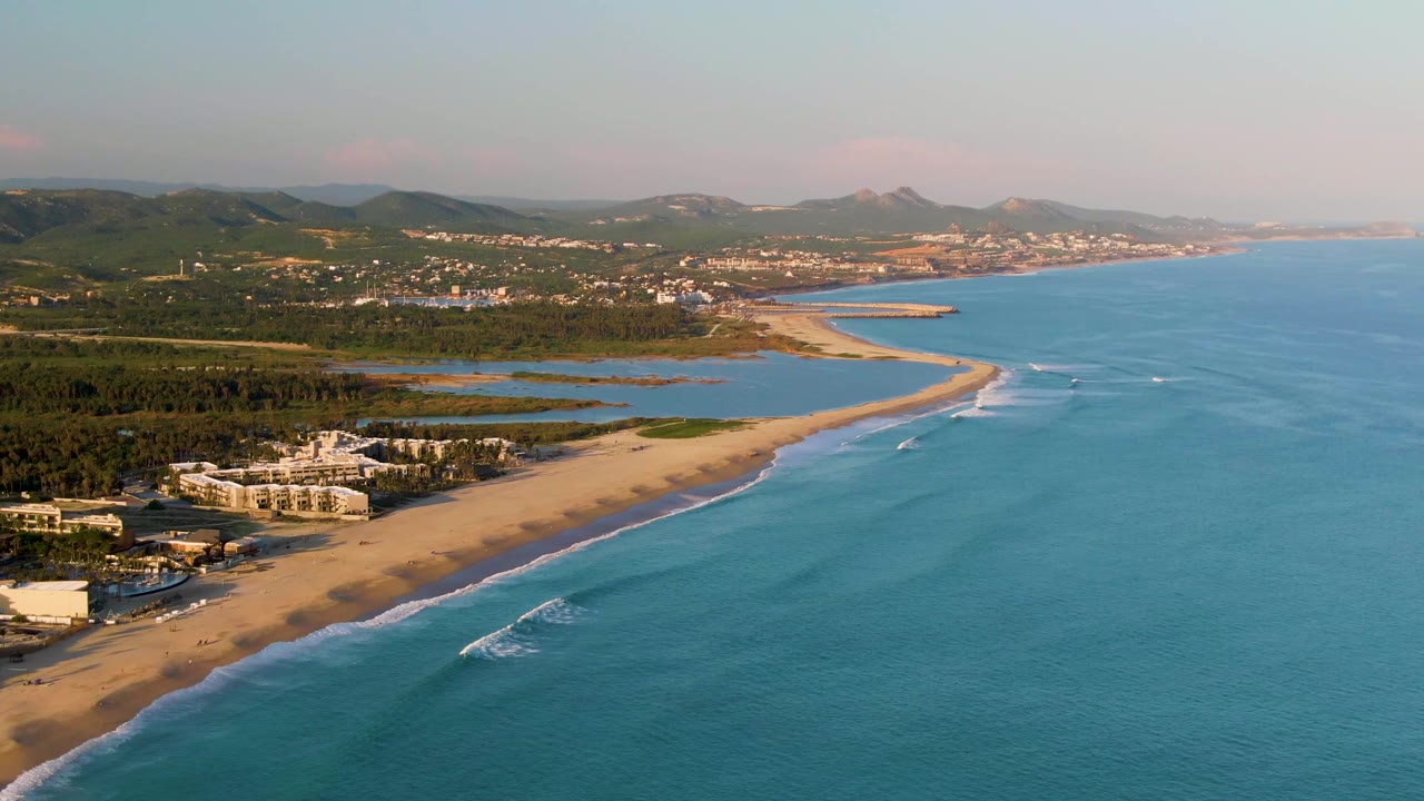 Aerial view over San Jose del Cabo coastline at 0:00, with turquoise ocean on the right, sandy beach and small white surf lines below, shoreline buildings to the left, distant hills, and no visible people.