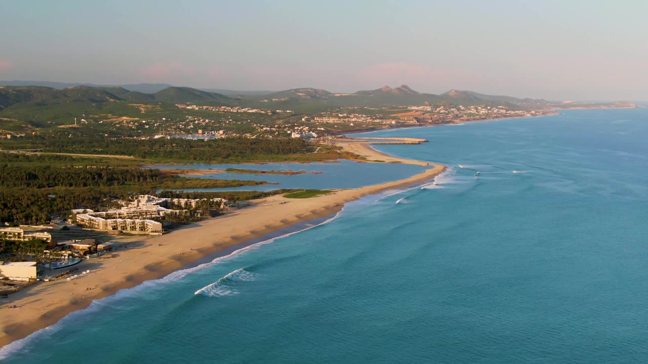 Drone view of ocean waves approaching a sandy beach beside coastal buildings.