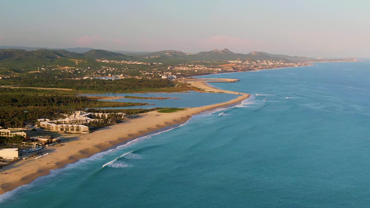 At 0:02, the aerial view shows San Jose del Cabo coastline with a broader stretch of blue water, white surf tracing the beach, low-rise buildings near the sand, green land behind them, and no visible people.