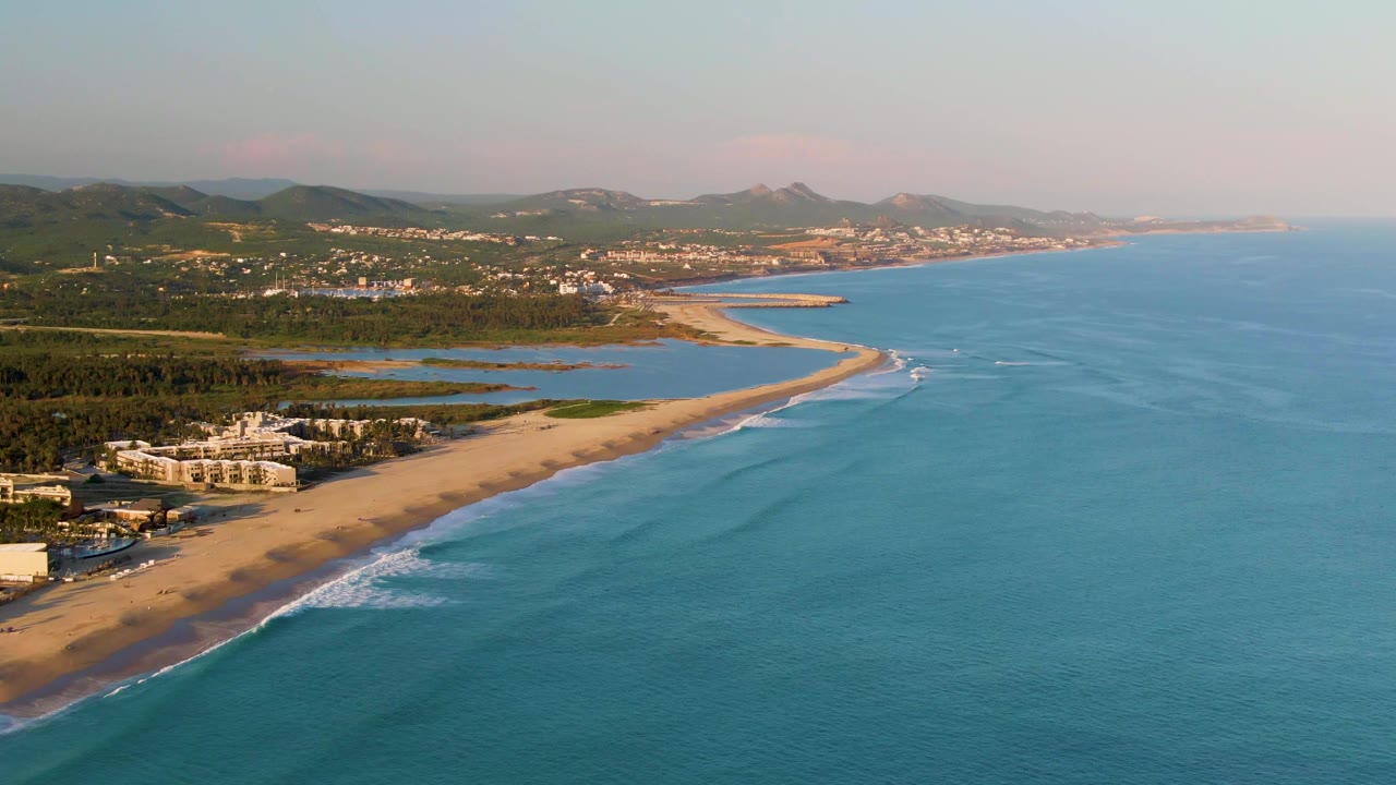 Aerial view of San Jose del Cabo coastline with blue ocean on the right, sandy beach on the left, resort buildings near shore, low hills beyond, and no visible people.