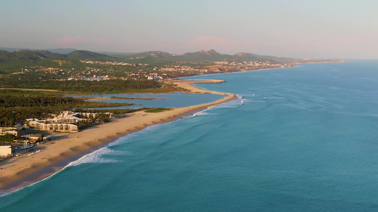 San Jose del Cabo aerial coastline view with a diagonal sandy shore, small white wave bands, calm blue water, scattered buildings, distant hills, and no visible people.