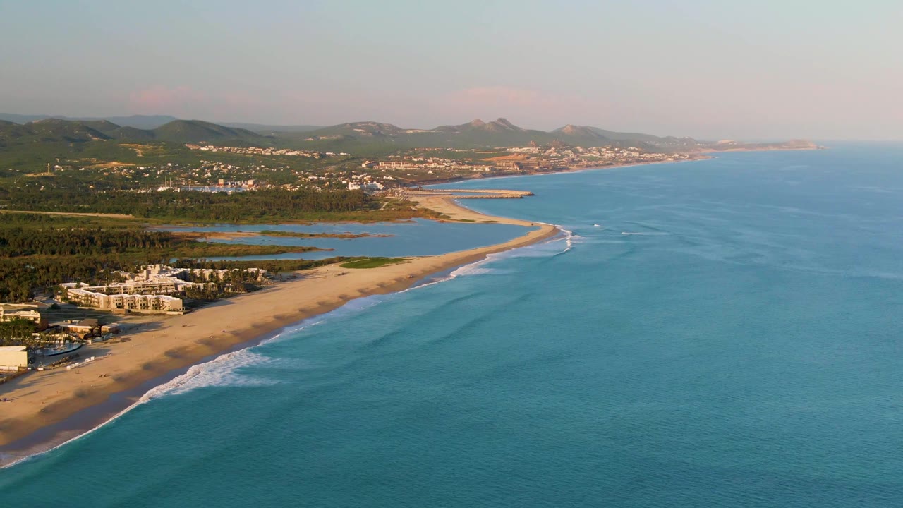Aerial frame over San Jose del Cabo showing more open blue sea to the right, a long sandy beach to the left, white surf near shore, low hills, buildings, and no visible people.