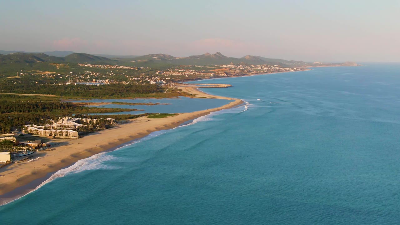 Coastal aerial with ocean foreground, sandy beach, lagoons, buildings, and distant hills.