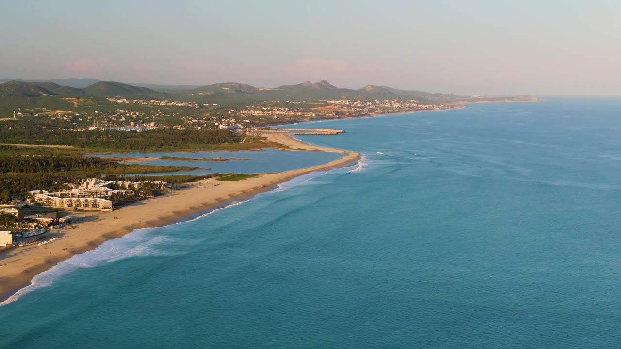 At 0:00, an aerial view of San Jose Del Cabo shows blue ocean in the foreground, a sandy beach curving past low buildings and green inland terrain, with no visible people.