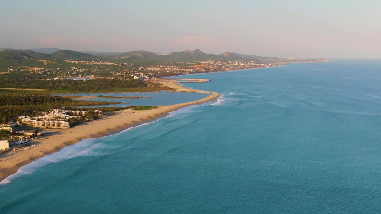 Drone-style coastal view with white foam along the beach and clear blue water offshore.