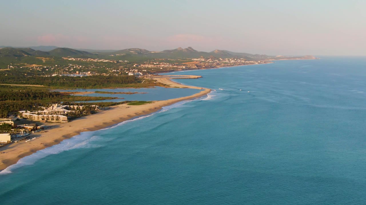 At 0:02, a wide aerial frame over San Jose Del Cabo shows more open water to the right, the beach and surf line to the left, and distant hills beyond coastal development, with no visible people.