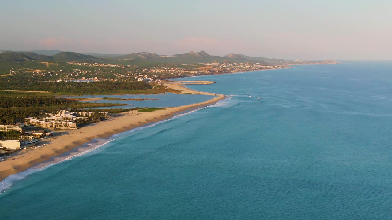Aerial opening frame over San Jose del Cabo coastline with turquoise ocean on the right, a sandy beach on the left, small boats near the shore, low buildings, lagoons, distant hills, and no visible people.
